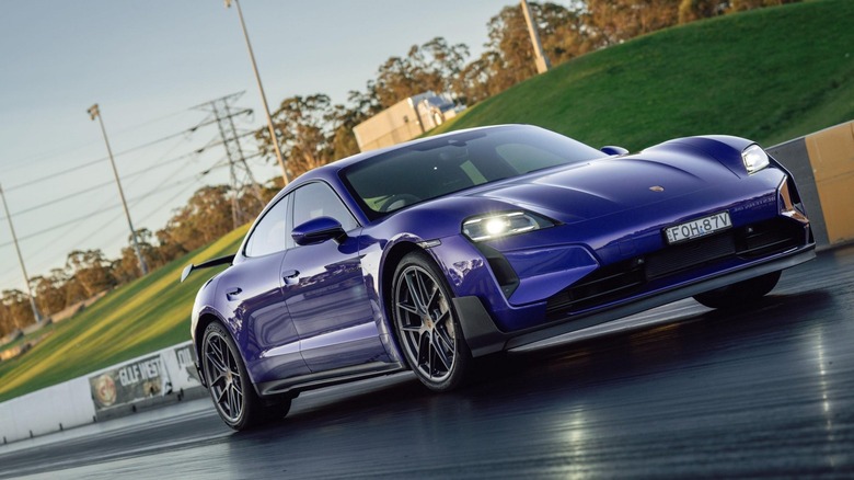 Low-angle front three-quarter shot of a metallic purple Porsche Taycan Turbo GT with Weissach Package on a race circuit with its headlights illuminated.