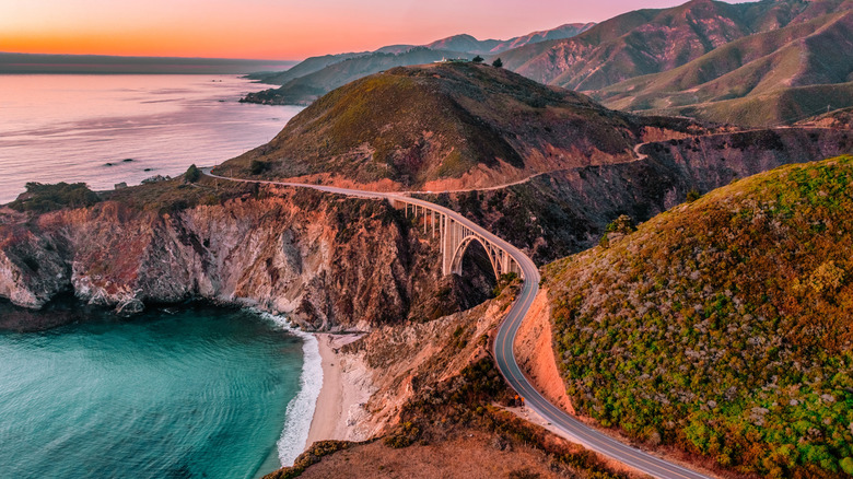 An overhead shot of the Pacific Coast Highway at the Bixby Bridge with the Pacific Ocean to the left and the rolling mountains to the right.