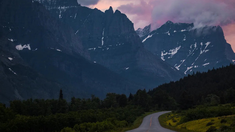 A road carved through the mountains, surrounded by green and trees, heading towards a mountain range with a purple and pink sunset behind them.