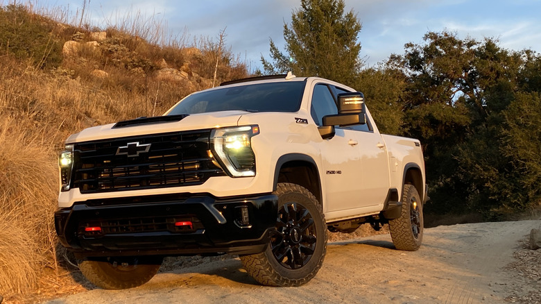 front three quarters shot of a white Chevy Silverado HD parked on dirt in front of dry brush and trees