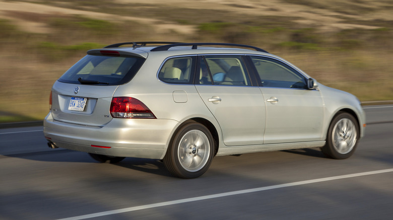 rear-side view of a light colored Jetta Sportwagen TDI driving next to a grassy hill