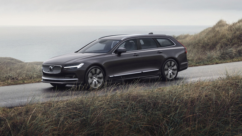 side view of a dark colored V90 driving on a coastal road with lots of grass in front of it