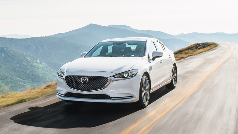 A front three-quarters shot of a white Mazda 6 driving on a cliffside road in front of a view of green mountains