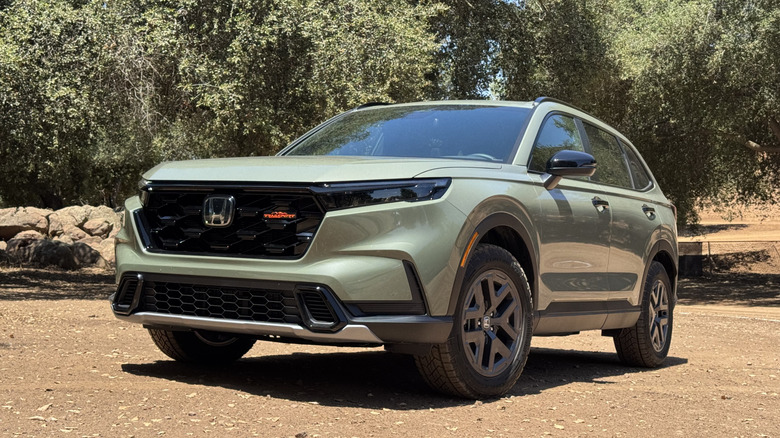 A front three quarters shot of a sage green Honda CR-V Trailsport parked on dirt in front of trees