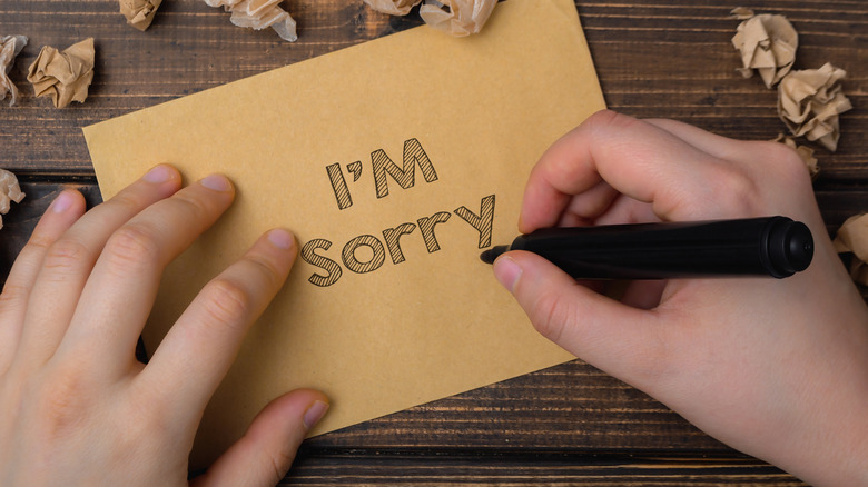Person is writing the word im sorry on a piece of paper. The paper is on a wooden surface and there are some shredded pieces of paper around it. The person is using a marker to write the word sorry
