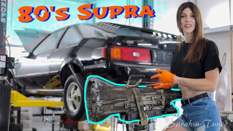 A black, '80s Supra on a car lift, with a transmission on a separate lift and Sarah in a black crop top tee and blue jean cutoffs.