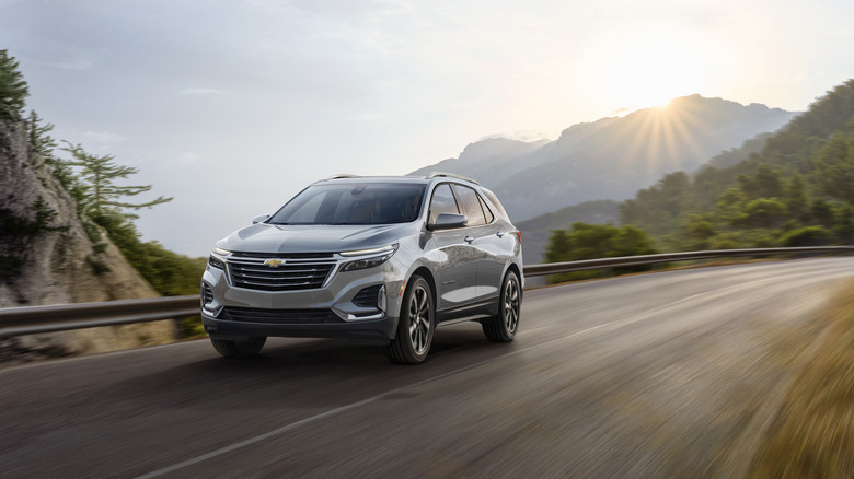 A silver, 2023 Chevrolet Equinox from the front 3/4 driver's side on a mountain road with the sun setting behind the mountains.