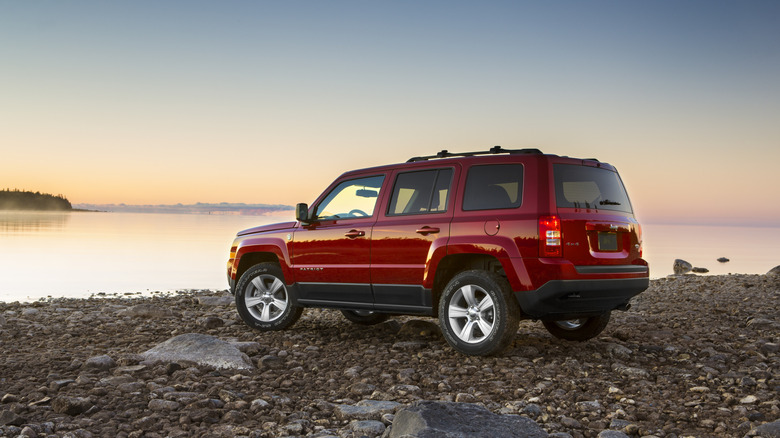 A red, boxy 2013 Jeep Patriot pondering its existence on a rocky beach beside a large body of water at sunset.