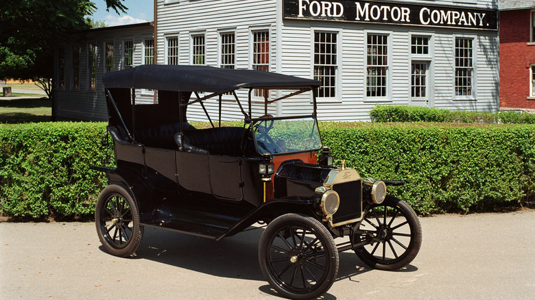A black, 1912 Ford Model T on a dirt road in front of a small white building with a sign that reads "Ford Motor Company."