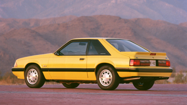 A yellow 1990 Ford Mustang LX on a rocky flat top set in the mountains.
