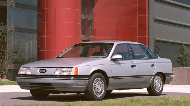 A silver, 1986 Ford Taurus on an asphalt driveway in front of a windowed building with red tiling.