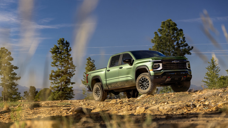 A green 2027 Chevrolet Silverado 1500 Trail Boss on a rocky terrain with a blue sky and sparse trees in the background.