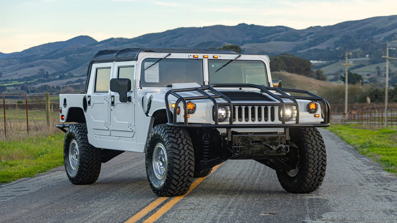 A white, obnoxiously large Hummer H1 parked on the center of a road across the double yellow lines in a rural area with mountains in the background.
