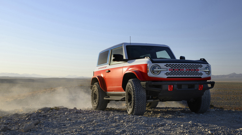The red, white and blue Bronco Stroppe edition sits on a rocky bed of road while the dust settles behind it under a blue sky with the mountains faintly seen on the horizon.