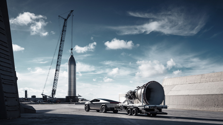 A Tesla Cybertruck doing Cybertruck things like just owing a space ship rocket booster on a trailer to a rocket staged in the background.