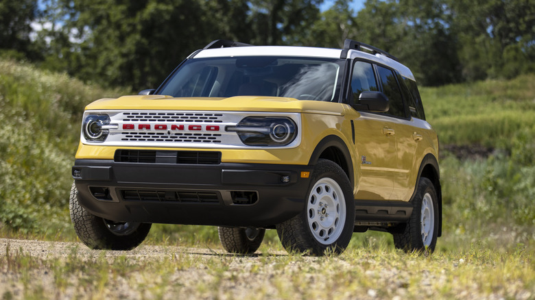 Front three quarters shot of a yellow and white Bronco Sport Heritage Edition parked on dirt in front of grassy hills