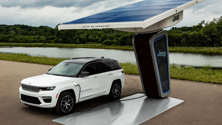 A white Grand Cherokee 4xe parked plugged into a Jeep solar powered charger in front of a lake