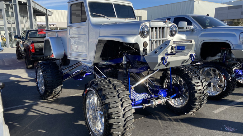 front three quarters shot of a vintage silver Jeep that's ridiculously lifted and chromed parked next to two other trucks