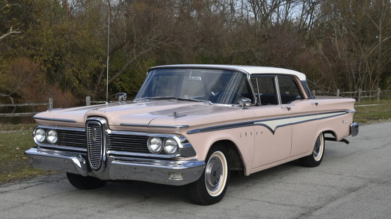 Front three quarters shot of a pink Edsel sedan parked on asphalt in front of lots of trees