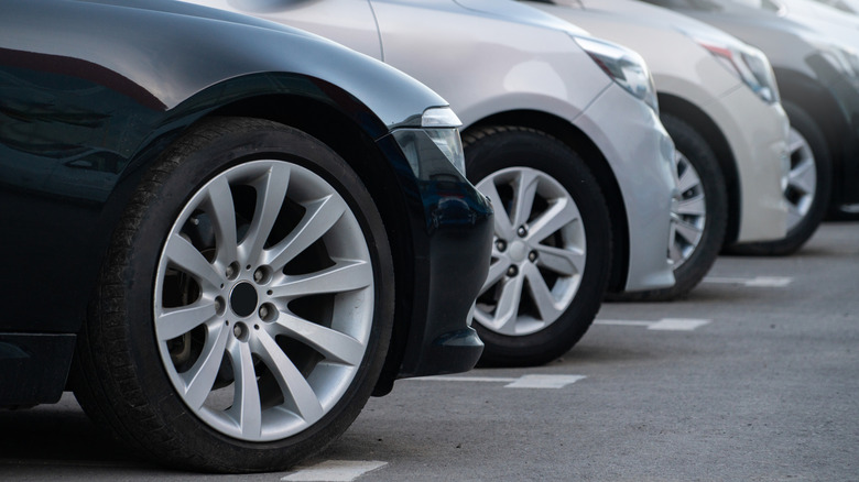 A row of car noses and the front passenger wheels, all in a grey, silver, beige color scheme.