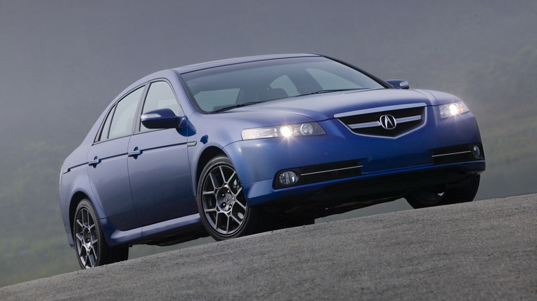 A front three-quarters shot of a blue Acura TL Type S with its headlights on parked on asphalt in front of a hazy background