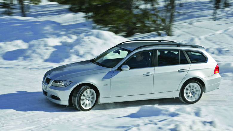 Side view of a silver BMW 330i wagon sliding on a snowy road in front of trees