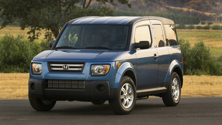 A front three-quarters shot of a blue Honda Element parked on blacktop in front of dead grass and some bushes and trees
