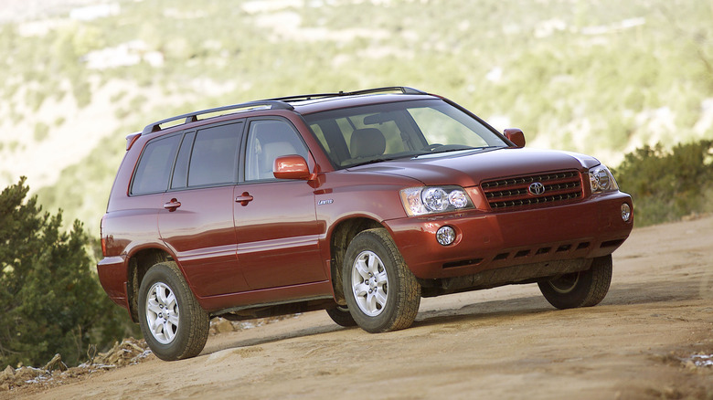 Front three quarters shot of a dark red first-gen Highlander parked on dirt in front of shrubs