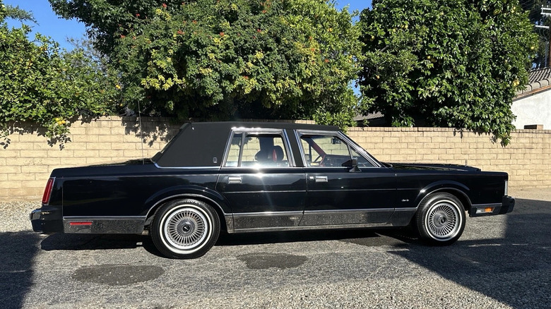 A black 1986 Lincoln Town Car with black sim top, sporting whitewall tires, parked in a driveway with a yellow brick backdrop.