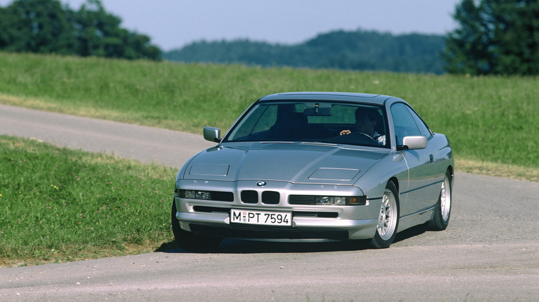 Front three quarters shot of a silver BMW 850i driving around a corner on a rural road flanked by grass