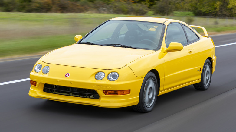 Front three quarters shot of a bright yellow Acura Integra Type R driving past trees and grass