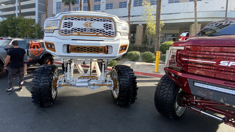 Photo of the front of a massive lifted white RAM HD next to a red Super Duty parked in front of palm trees and a building, dwarfing a tiny man standing next to them