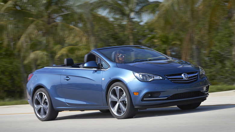 Front three-quarters image of a blue Buick Cascada being driven past a bunch of palm trees with its top down