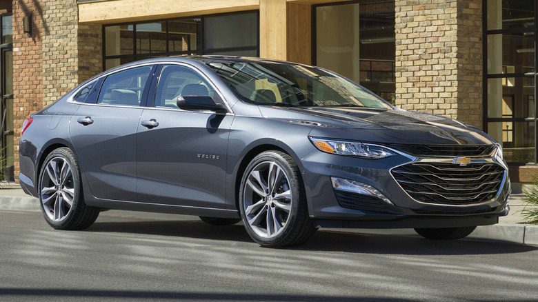 Front three quarters image of a dark gray Malibu parked on the street in front of a building