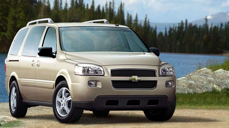 Front three-quarters image of a metallic beige Chevrolet Uplander parked on sand in front of a lake with trees and a blue sky in the background