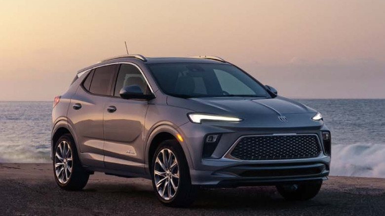 Front three-quarters image of a silver Buick Encore parked on the sand at the beach at dusk with waves crashing behind it