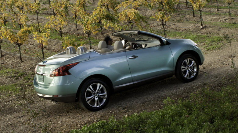 Rear three-quarters image of a light blue Murano CrossCabriolet with the top down parked on dirt in front of grapevines