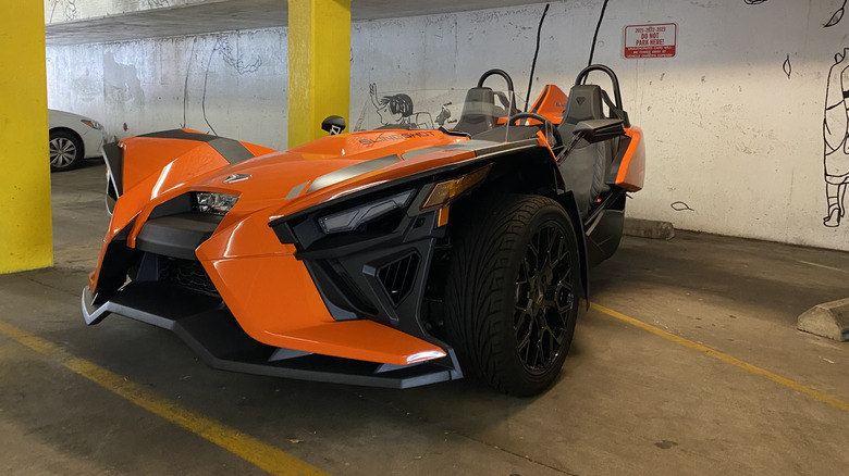 Front three-quarters image of an orange Polaris Slingshot parked in a garage next to yellow poles and a white wall