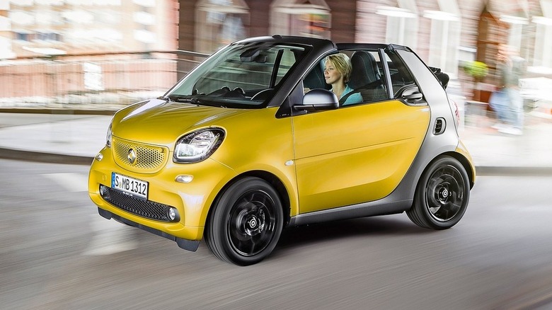 Front three-quarters image of a metallic yellow Smart ForTwo Cabriolet driving past blurred buildings