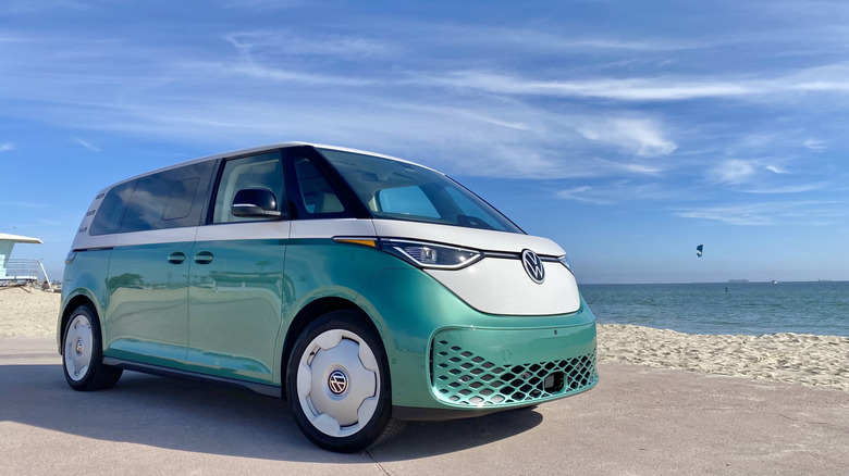 Front three-quarters image of a green-and-white ID Buzz parked on the beach with a cloudy blue sky and the ocean visible behind it