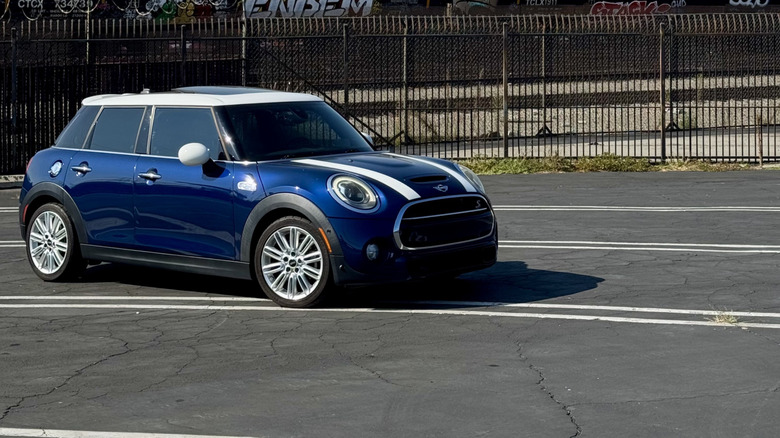 A front three quarters shot of my blue 2017 Mini Cooper S four-door parked in a lot in front of a fence, train tankers, and the downtown LA skyline