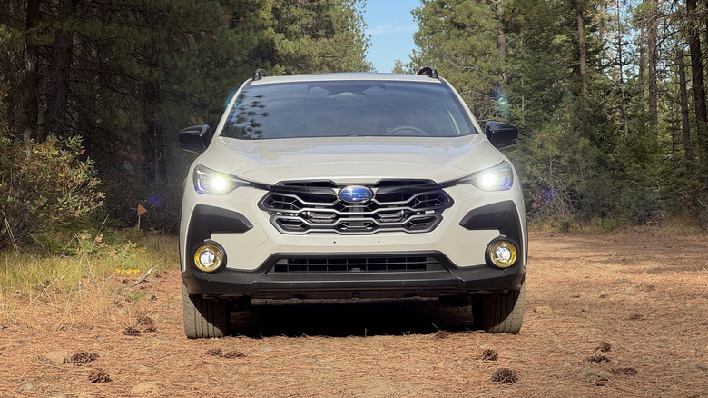 A head-on shot of a white 2026 Subaru Crosstrek Hybrid parked on dirt with its headlights on in the forest in front of trees and a blue sky