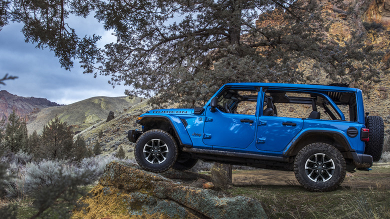 A side view of an electric blue Jeep Wrangler Unlimited with its front wheel on a rock in front of mountains and trees