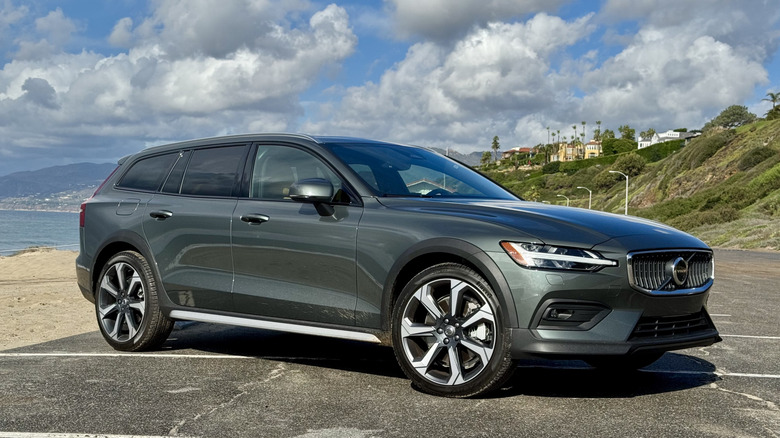 Front three quarters shot of a dark green 2026 Volvo V60 Cross Country parked in a beachside parking lot in front of sand, clouds, and a green cliff