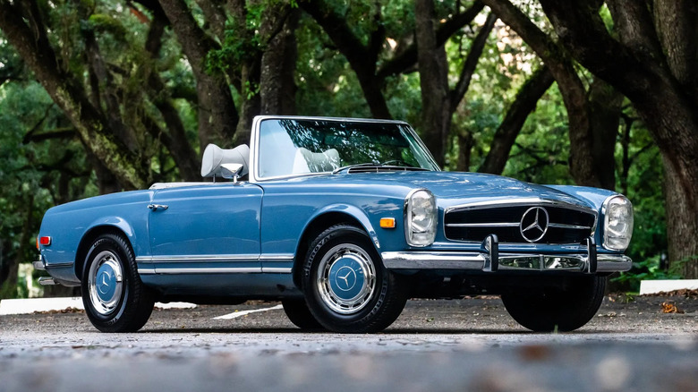 Front three quarters shot of a light blue Mercedes 280 SL with the top down parked in a lot in front of dense trees