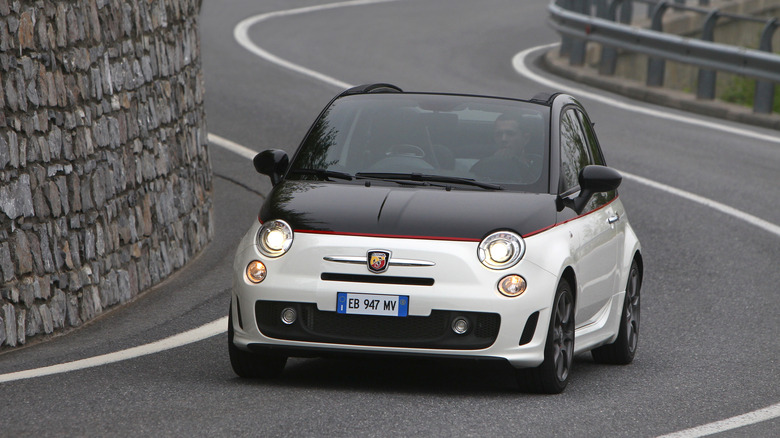 Front three quarters shot of a black and white Fiat 500 C Abarth driving on a winding road next to a brick wall