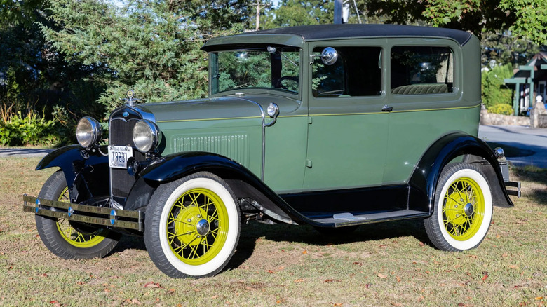 Front three quarters shot of a green Ford Model A with yellow wire wheels parked on grass in front of trees