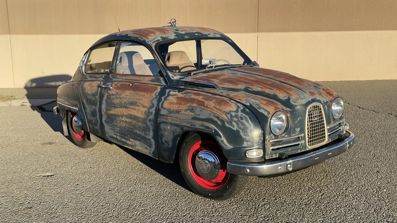 A front three quarters shot of a patina'd '64 Saab 96 parked on asphalt inf ront of a tan wall