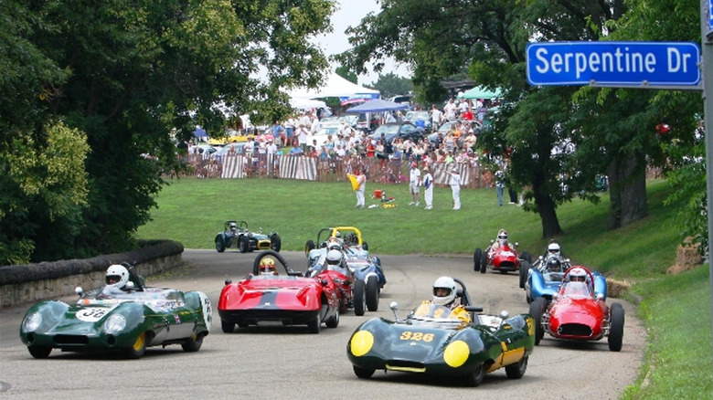 A photo of a gaggle of vintage cars racing at the PVGP in front of grass and a sign for Serpentine Dr