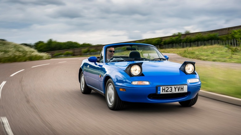 A blue NA Miata driving around a corner with its headlights up and roof down next to grass and bushes